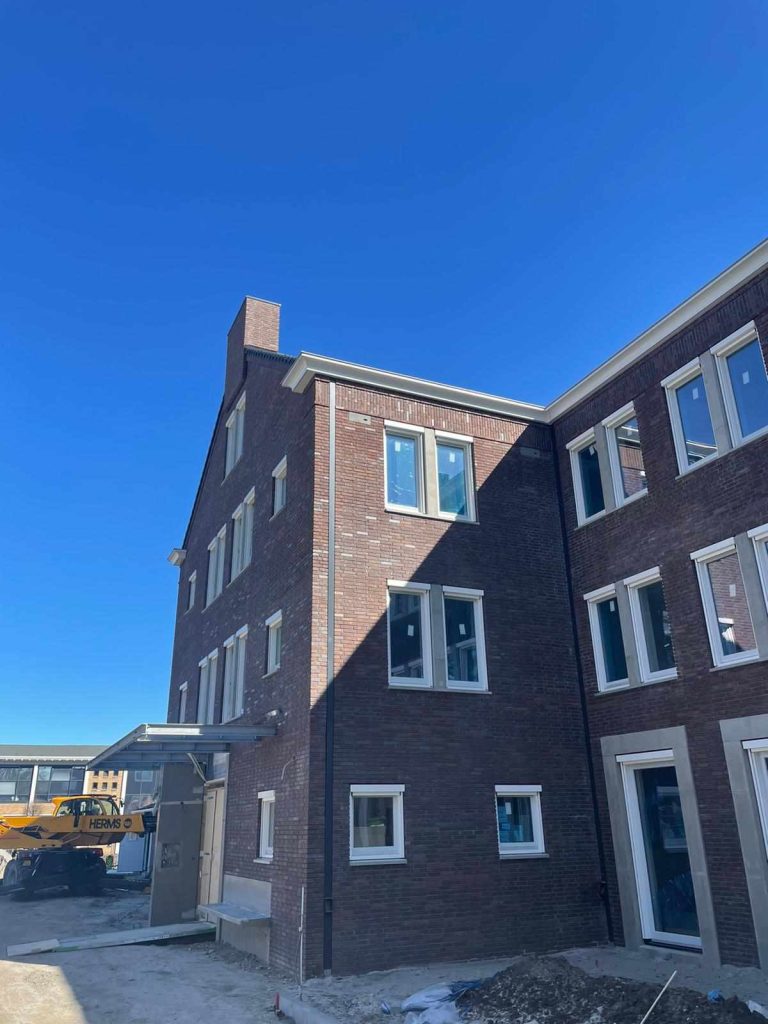 Hoek van het gebouw in donkerrode baksteen met een schoorsteen en een luifel boven de ingang