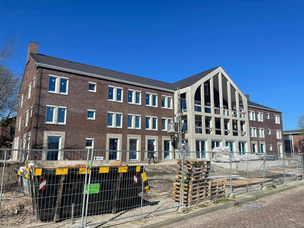 Het gebouw in de afbouwfase: donkere baksteen, een betonnen puntgevel met balkons en bouwhekken op de voorgrond